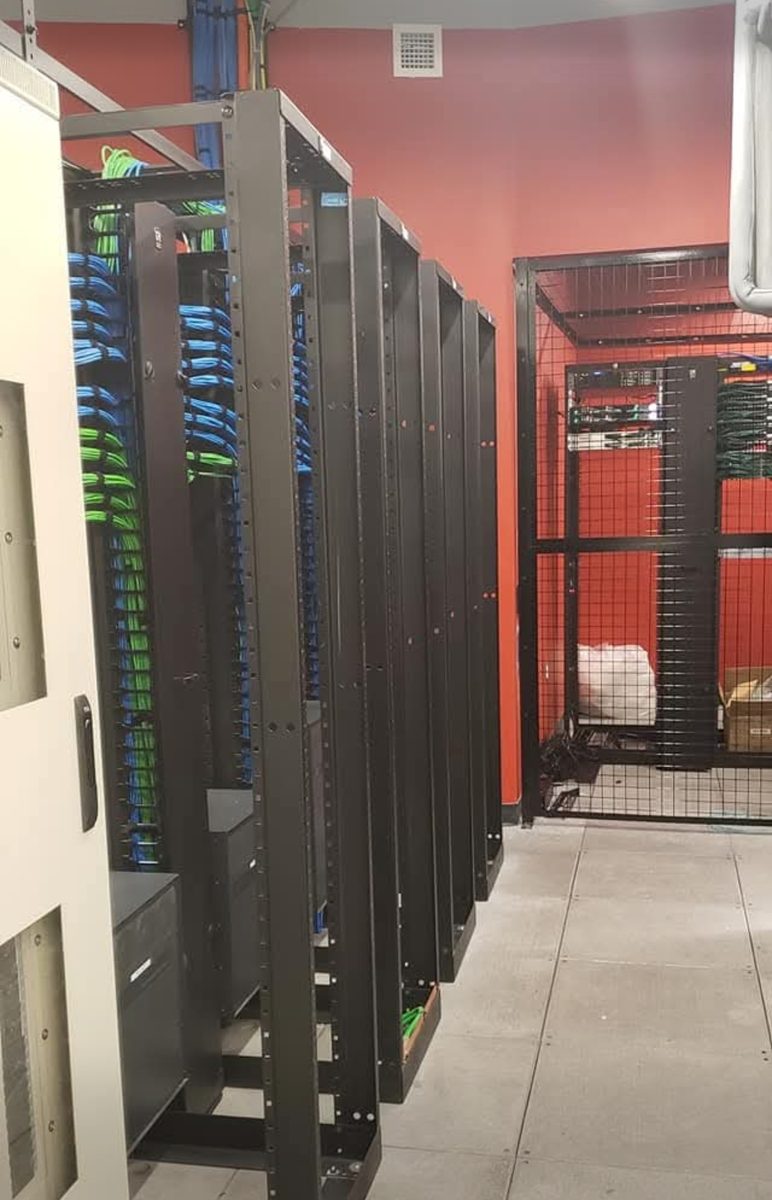 Row of open-frame network racks in a data center equipment room with blue and green cabling terminated to patch panels, installed by DataTel 360