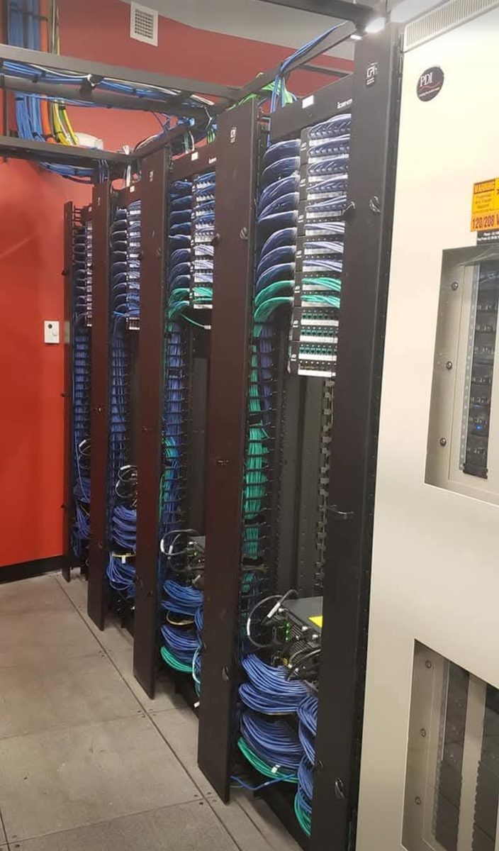 Enterprise data center racks loaded with terminated copper patch panels, fiber panels and colour-coded patch cabling beside a power distribution unit, installed by DataTel 360