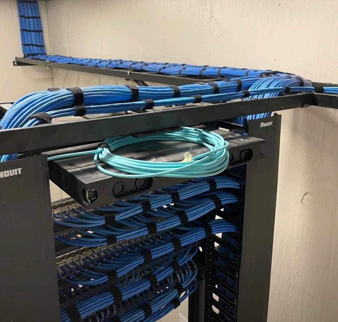 Blue Cat6 cabling dressed and secured across a ladder rack cable runway above a network rack, with a fiber patch cord coiled below, installed by DataTel 360